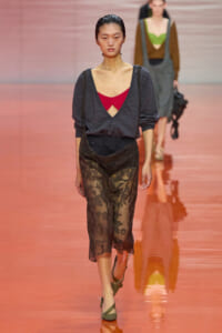 Model on a runway wearing a dark gray V-neck top over a red bralette and sheer black lace pants with green sandals.