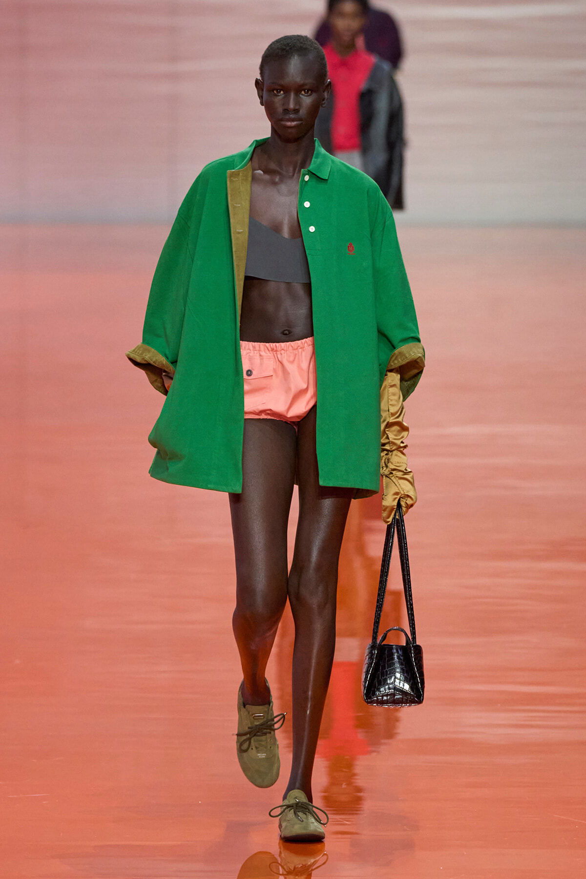 Runway model wearing a bright green oversized coat over peach shorts, gray top, tan gloves, and a small black handbag.