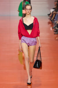 Model walking the runway in a red open-front blouse over a black bralette with lavender shorts, carrying a black handbag.