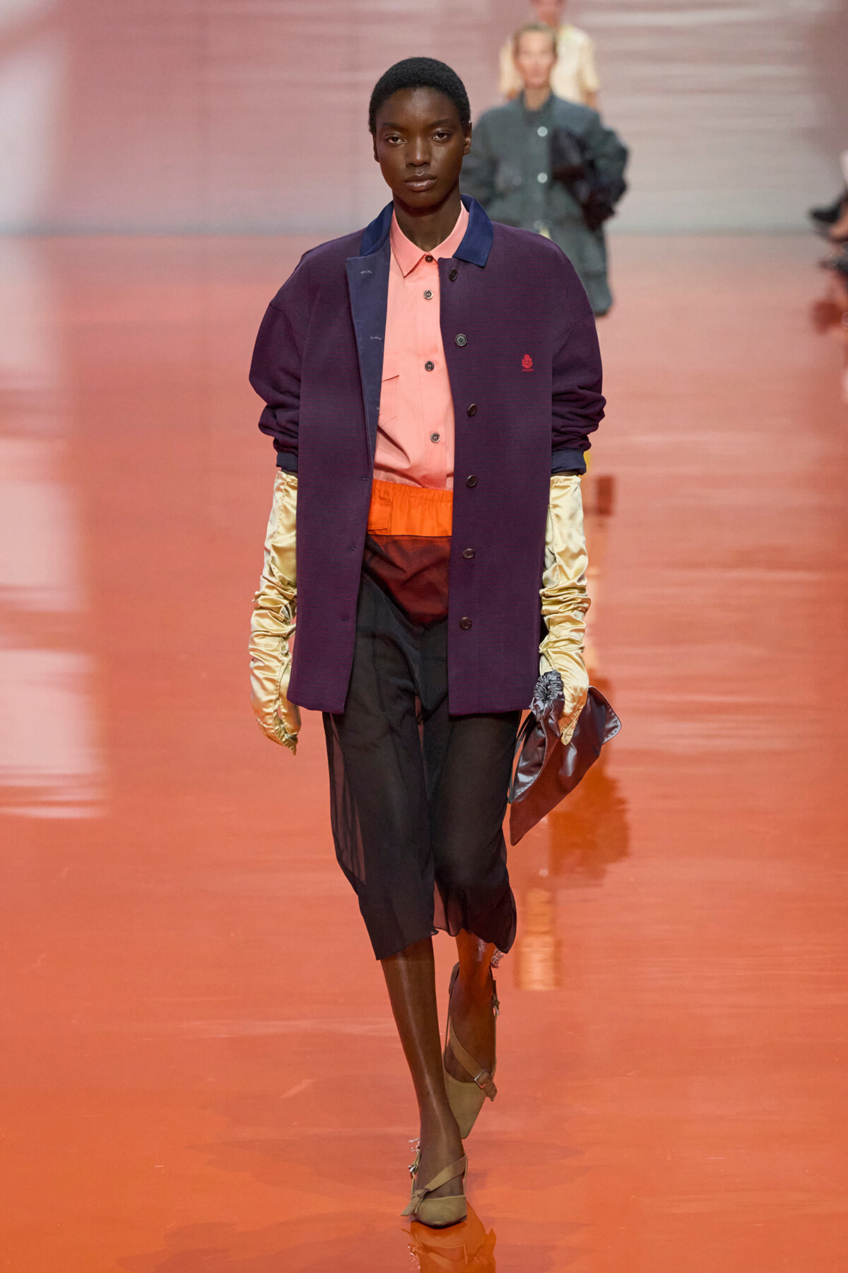 Model walks a fashion show in a peach shirt under a purple jacket with a navy collar, orange waistband, black cropped pants, and gold satin sleeves, carrying a metallic clutch.