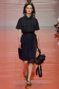 Model walking the runway in a black short-sleeve shirt and navy belted skirt, carrying a black bag with straps.