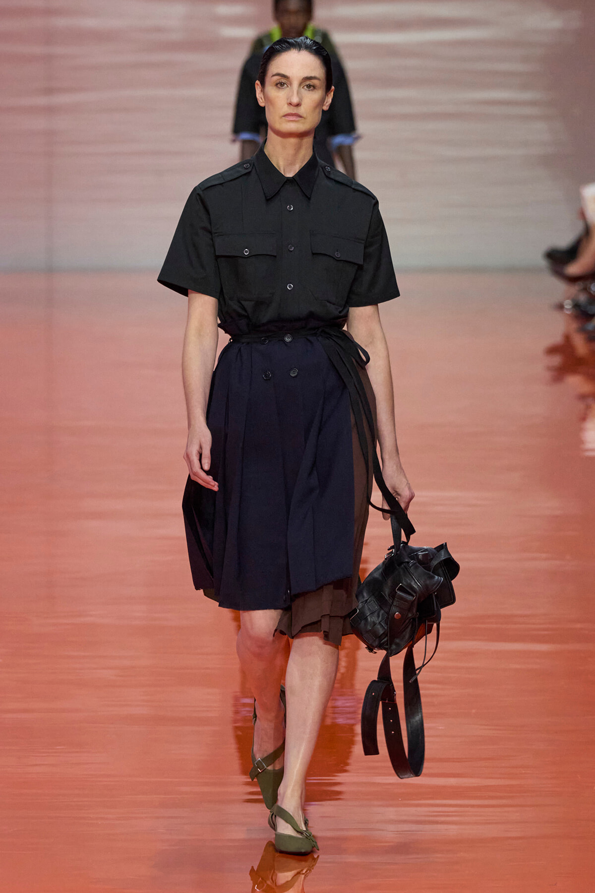 Model walking the runway in a black short-sleeve shirt and navy belted skirt, carrying a black bag with straps.