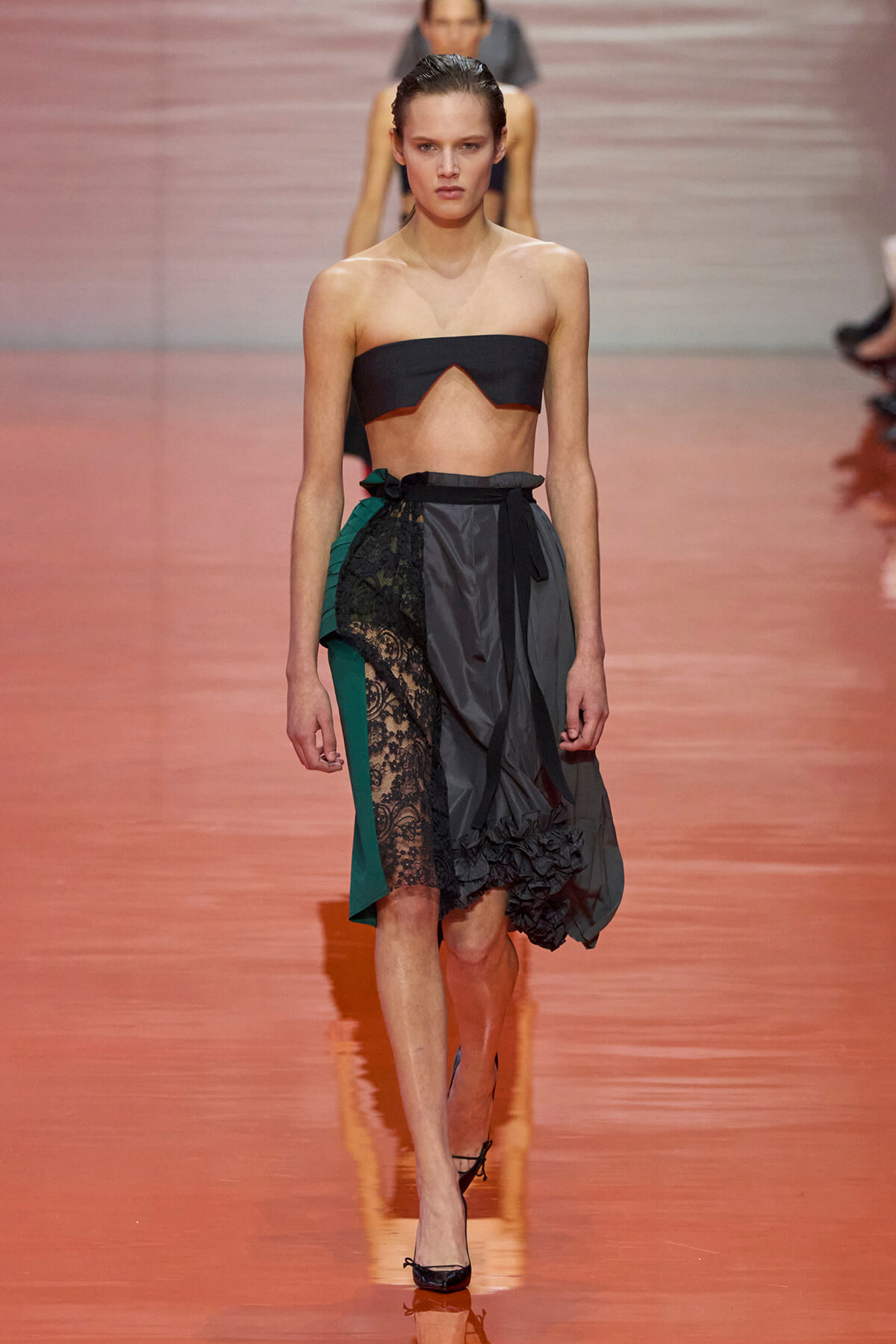 Model on a runway wearing a strapless black bandeau top and a color-blocked midi skirt with lace, green and gray panels, tied at the waist, black heels.