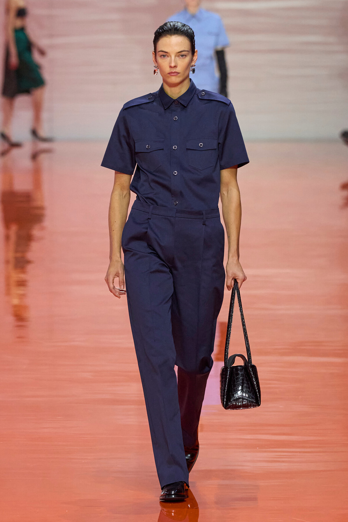 Model walks a runway in a navy utility jumpsuit with short sleeves, button front, two chest pockets, and a matching belt, carrying a small black croc-embossed handbag.