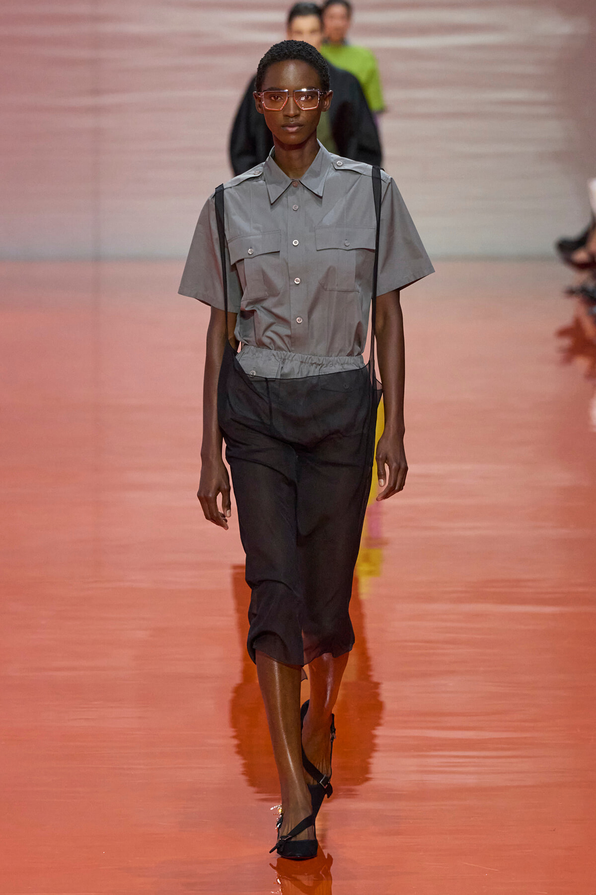 Model walks a runway in a gray short-sleeve button-up shirt with suspenders and black cropped pants, wearing orange glasses.