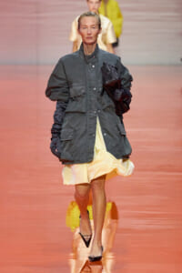 Model walks the runway in an oversized gray coat over a pale yellow dress, clutching a black bag and wearing black heels.