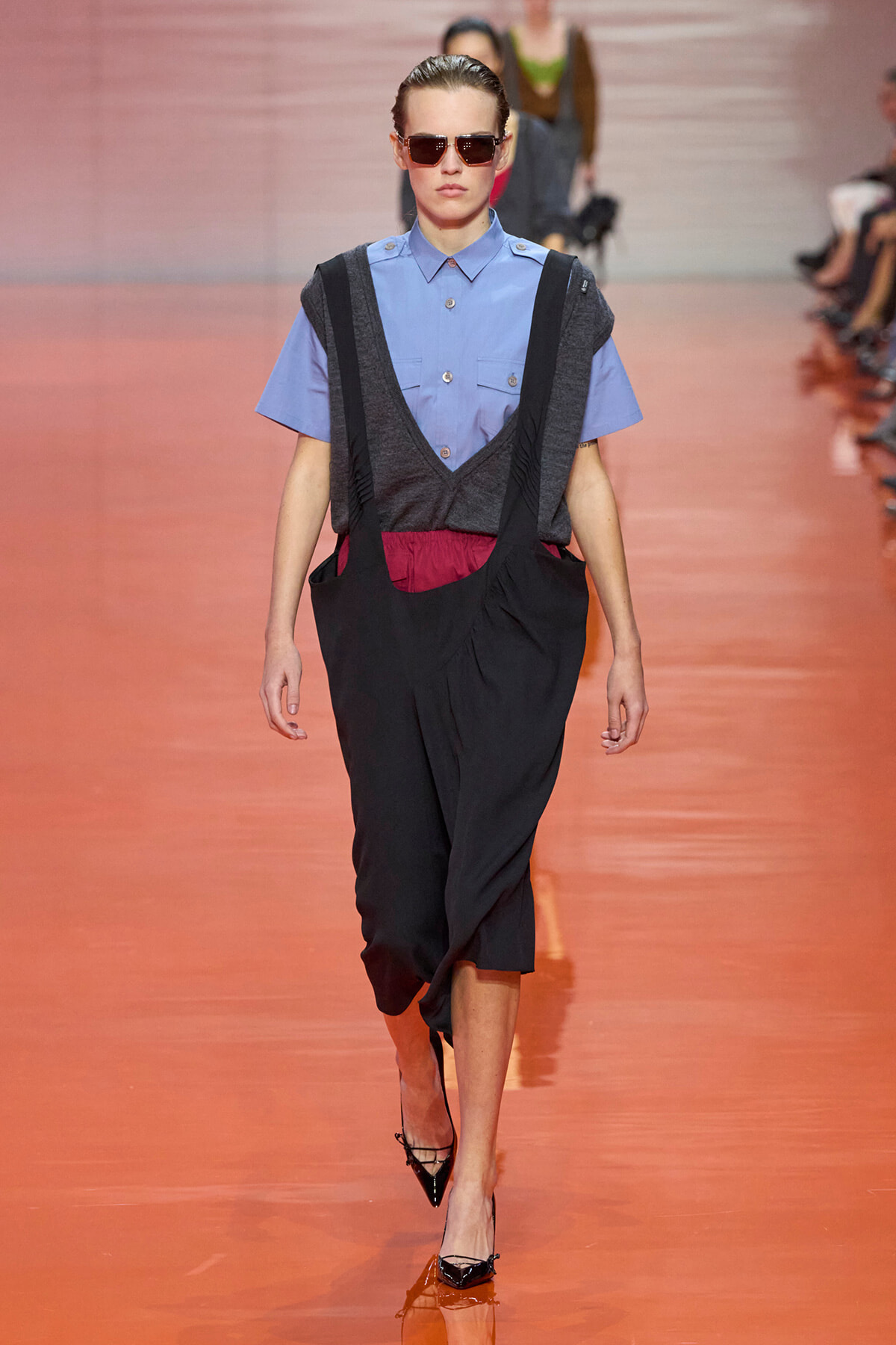 Runway model wearing a light blue short-sleeve shirt with a gray layered vest and black suspender-style pants, red waistband, sunglasses, walking down the catwalk.