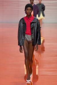 Model walks a pink runway wearing a red button-down shirt under an oversized black leather jacket and light gray shorts, with white flats visible.