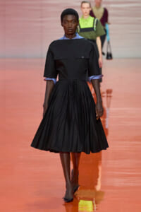 Model walking a runway in a black knee-length dress with a fitted bodice and full skirt; blue shirt cuffs peek from rolled sleeves.