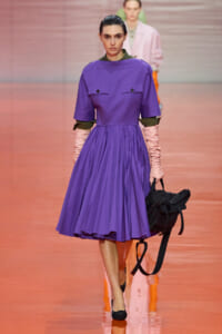 Model on a runway wearing a knee-length purple dress with structured bodice, green cuffs, pink gloves, and a black backpack-like bag.