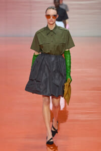 Model walking runway in olive utility shirt, full gray A-line skirt, green gloves, brown sunglasses, and tan clutch.