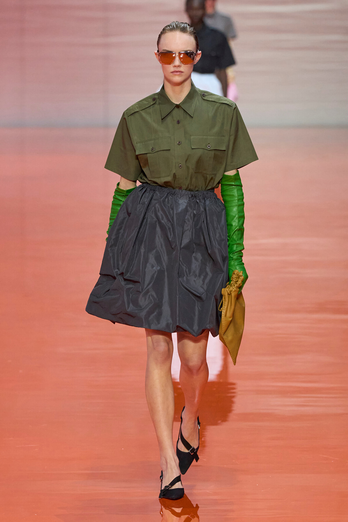 Model walking runway in olive utility shirt, full gray A-line skirt, green gloves, brown sunglasses, and tan clutch.