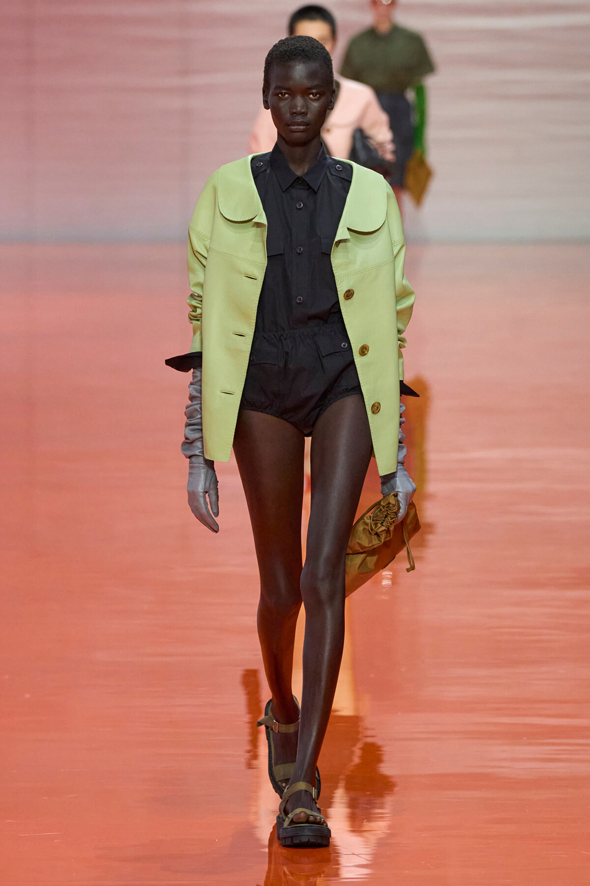 Model walking a fashion runway in a lime oversized jacket over a black shirt and shorts, with gray gloves and sandals.