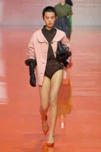 Model walking a runway in a pale pink oversized coat over a dark bodysuit, with black gloves and orange pointed heels, carrying a black handbag.