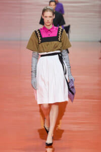 Model walking the runway in a pink-caced collar top under an olive-brown overlay with embellished straps, paired with a white pleated skirt, gray gloves, a black belt, and a lavender clutch.