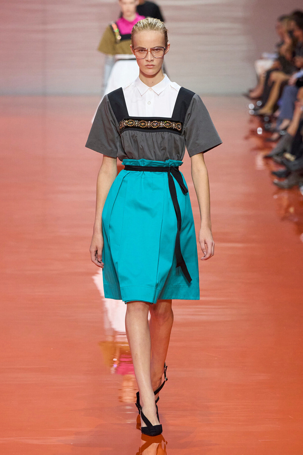 Model on a runway wearing a teal knee-length skirt with a black belt, paired with a gray short-sleeve top featuring a white collar and jeweled band, and pink-tinted glasses.