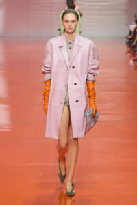 Model walking a fashion show in a pastel pink knee-length coat with rolled sleeves, wearing a gray-green outfit underneath and bright orange gloves, carrying a gray clutch.