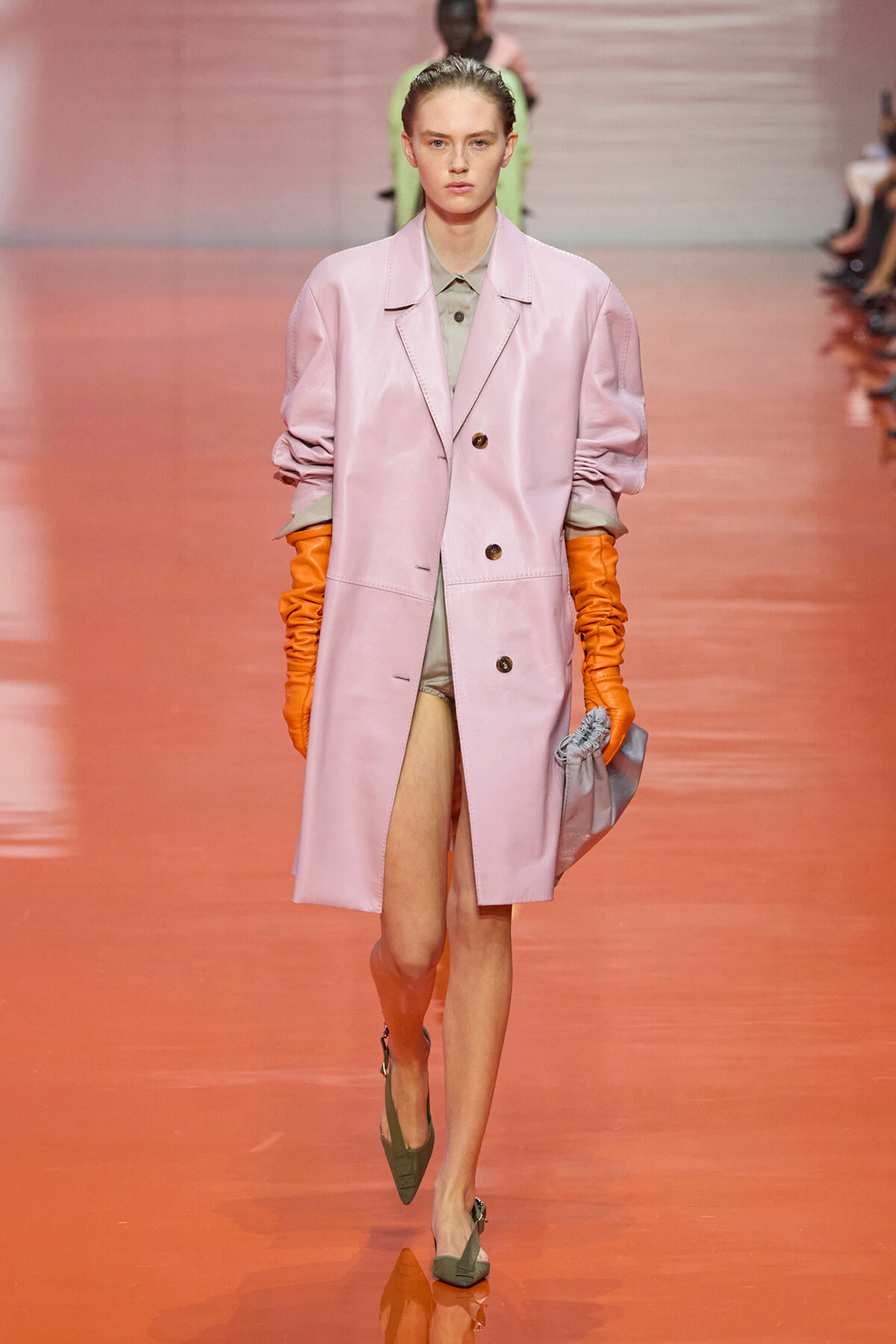 Model walking a fashion show in a pastel pink knee-length coat with rolled sleeves, wearing a gray-green outfit underneath and bright orange gloves, carrying a gray clutch.