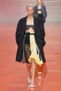 Model walking the runway in a black oversized coat over a color-block dress with lace panel and a yellow ruffled hem, white heels visible.