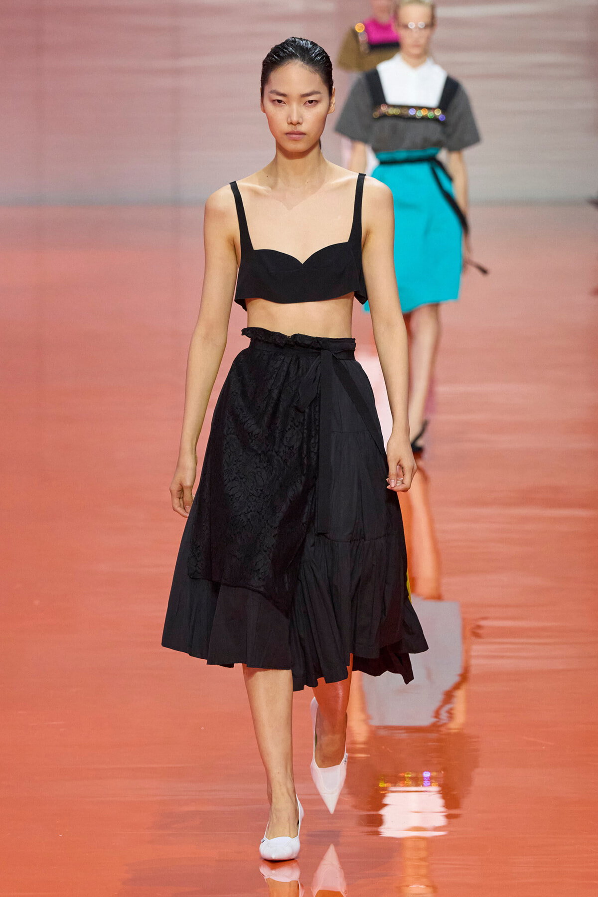 Model walks the runway wearing a black bralette-style top and a high-waisted, tiered black midi skirt, paired with white flats on a peach-colored catwalk.