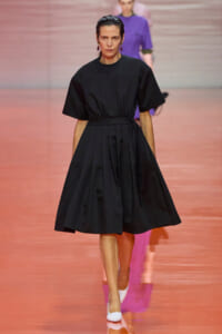 A woman models a knee-length black dress with a wide belt on a pink runway, white shoes, and sleek hair.