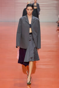 Runway model wearing an oversized gray blazer with a matching gray pleated midi skirt and olive heels, walking the catwalk in a fashion show.