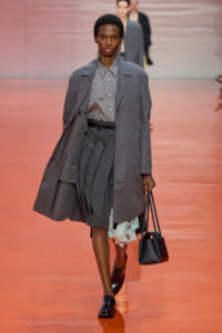 Model walking runway in gray ensemble: oversized trench coat over button-up shirt and pleated skirt, carrying a black handbag on a coral stage.