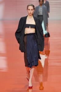 Model walks a fashion runway in a black oversized jacket, black bandeau top, and navy layered skirt, with red heels.