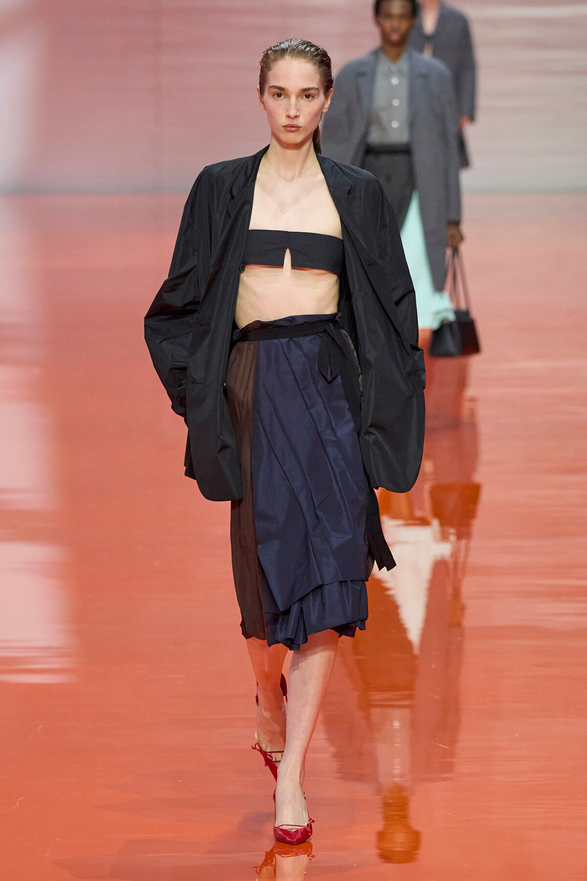 Model walks a fashion runway in a black oversized jacket, black bandeau top, and navy layered skirt, with red heels.
