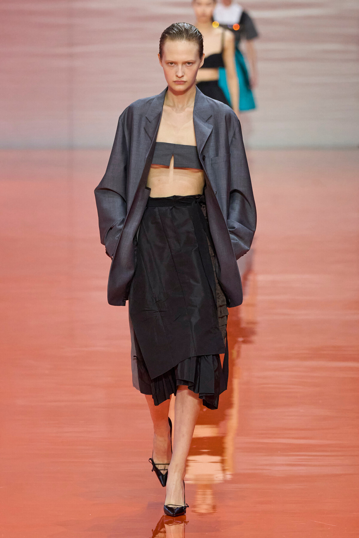 Model walking the runway in a loose gray blazer over a black bandeau and layered black skirt on a pinkish glossy catwalk.