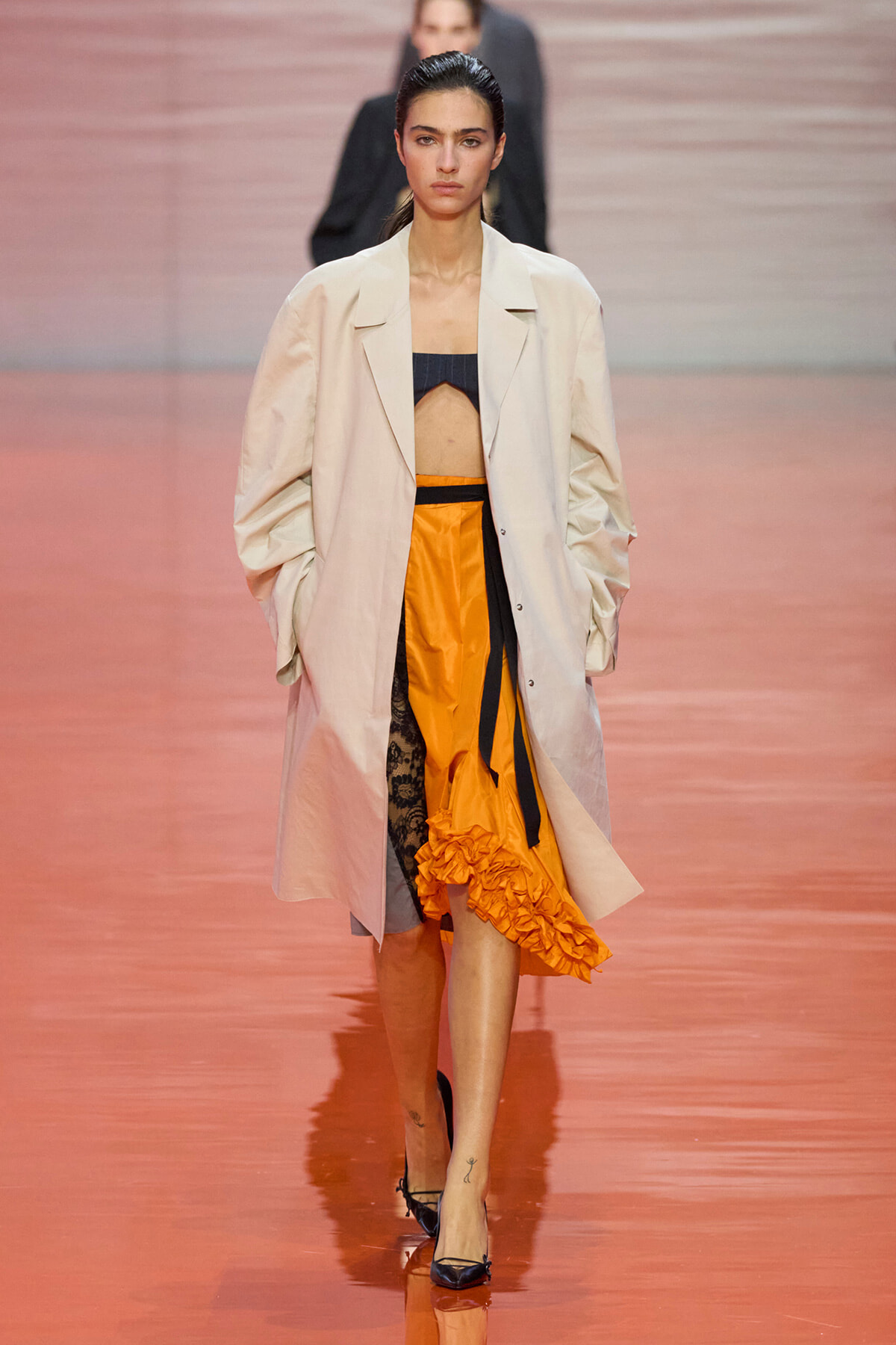 Model walking a runway in a pale beige oversized trench over an orange ruffled skirt and black crop top, with heels, on a pinkish runway.