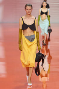 Model walking the runway in a yellow draped dress with a black cropped top, long mustard gloves, and a black handbag.