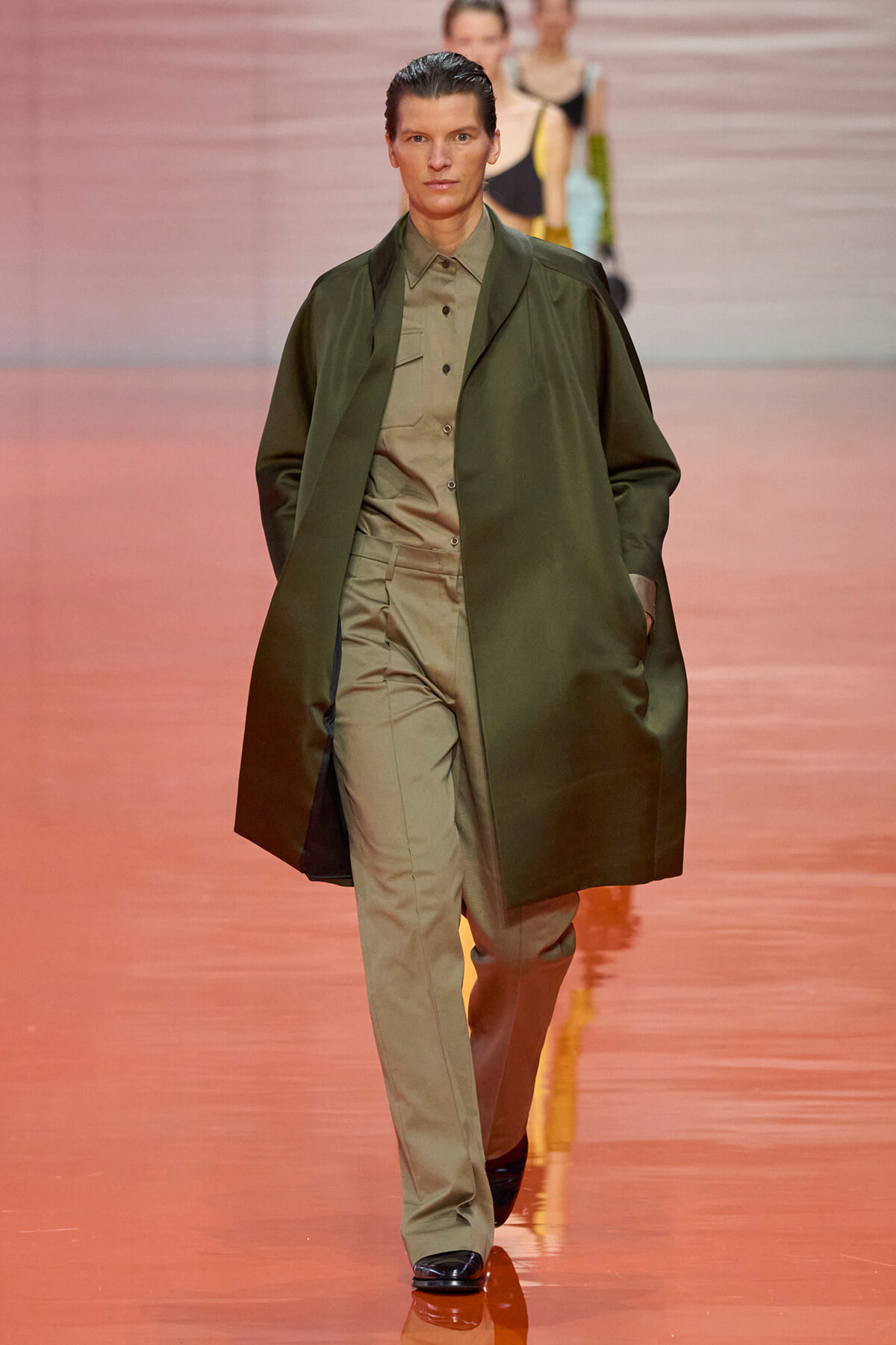 Model walks the runway wearing an olive-green long coat over a khaki suit during a fashion show.