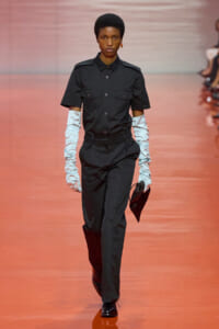 Model walks a fashion runway in a black short-sleeve utility shirt with epaulets, black trousers, white satin gloves, and a black clutch.