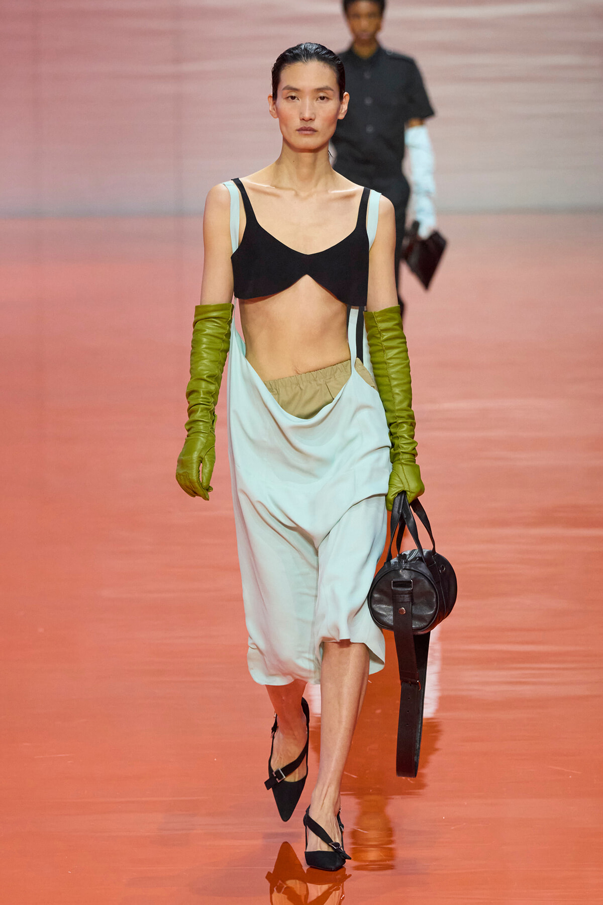 Model walking a runway in a black crop top over pale blue draped pants, olive-green gloves, and a small round black handbag.