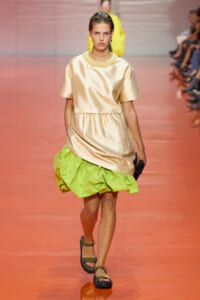 Model walking a fashion runway in a pale gold satin tunic with a lime-green puffed skirt and sandals.