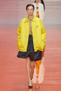 Model walking a runway in a bright yellow oversized jacket over a tan button-down shirt and a dark gray voluminous skirt, wearing pink-tinted glasses with black heels.