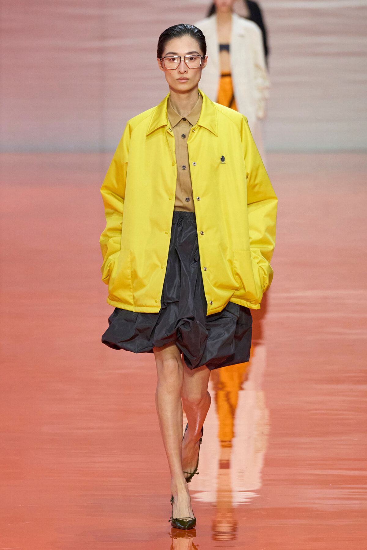 Model walking a runway in a bright yellow oversized jacket over a tan button-down shirt and a dark gray voluminous skirt, wearing pink-tinted glasses with black heels.