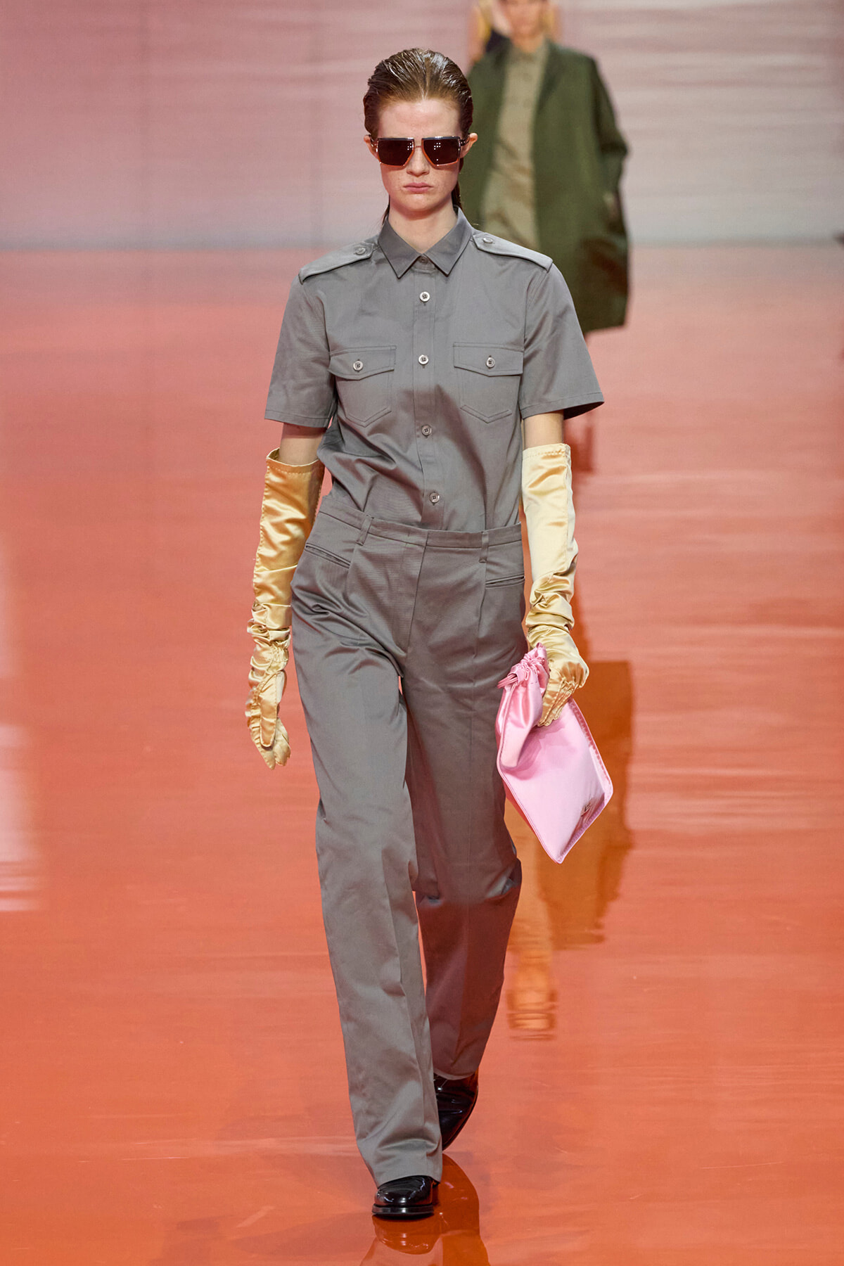 Female model walks a fashion runway in a gray utility jumpsuit, short sleeves, sunglasses, satin gloves, and a pink clutch.