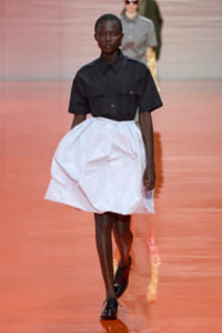 Model on a runway wearing a black short-sleeve button-up shirt and a voluminous white knee-length skirt with black shoes.