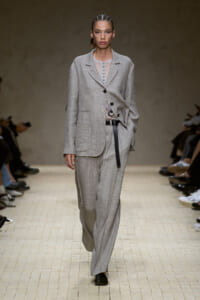 Model walks the runway in a light gray oversized suit: single-breasted blazer with large pockets and a thin belt over matching wide-leg pants.