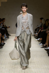 Runway model in a light grey blazer over a draped top and wide, patterned trousers, carrying a beige woven tote.