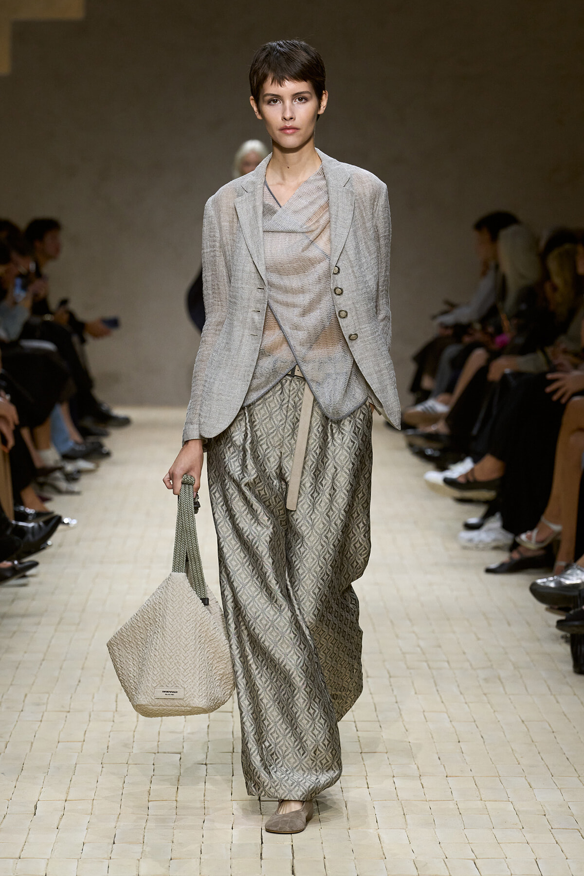 Runway model in a light grey blazer over a draped top and wide, patterned trousers, carrying a beige woven tote.