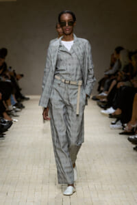 Model walking the runway in a light gray plaid suit with matching vest, oversized jacket, beige belt, and white loafers, wearing dark sunglasses.