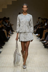 Model walks a fashion runway in a pale grey, patterned two-piece outfit (boxy jacket and pleated mini skirt) carrying a light grey fringe handbag on a beige tiled catwalk.