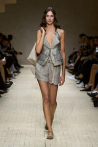 Runway model wearing a geometric gray vest over light gray shorts, carrying a large beige tote on her shoulder.