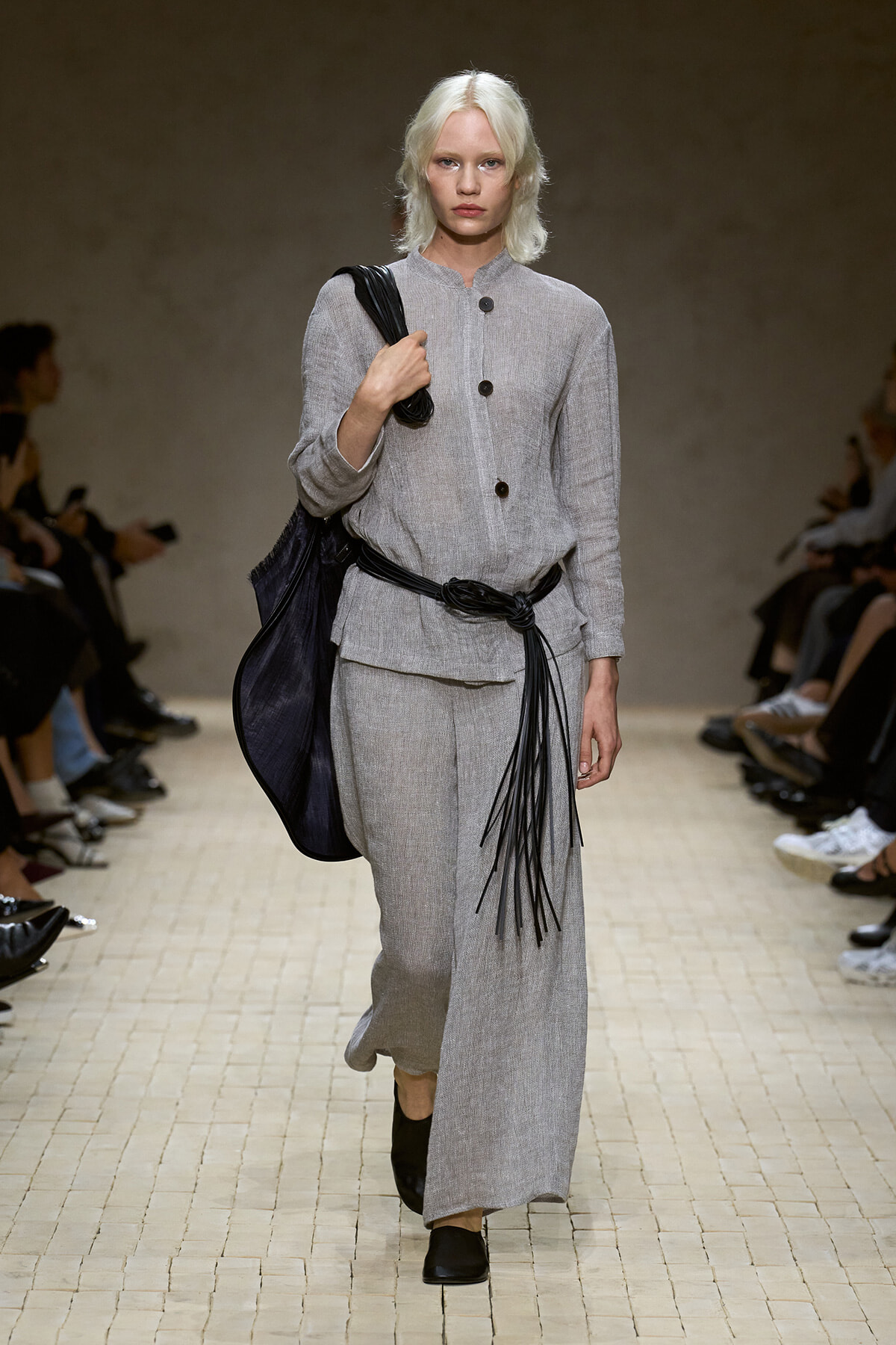 Model on a runway wearing a light gray linen matching top and wide-leg pants, with a large black fringe belt and a black shoulder bag.