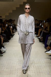 Model on a fashion runway wearing a pale gray tunic and wide-leg pants, round sunglasses, and a small silver circular crossbody bag.