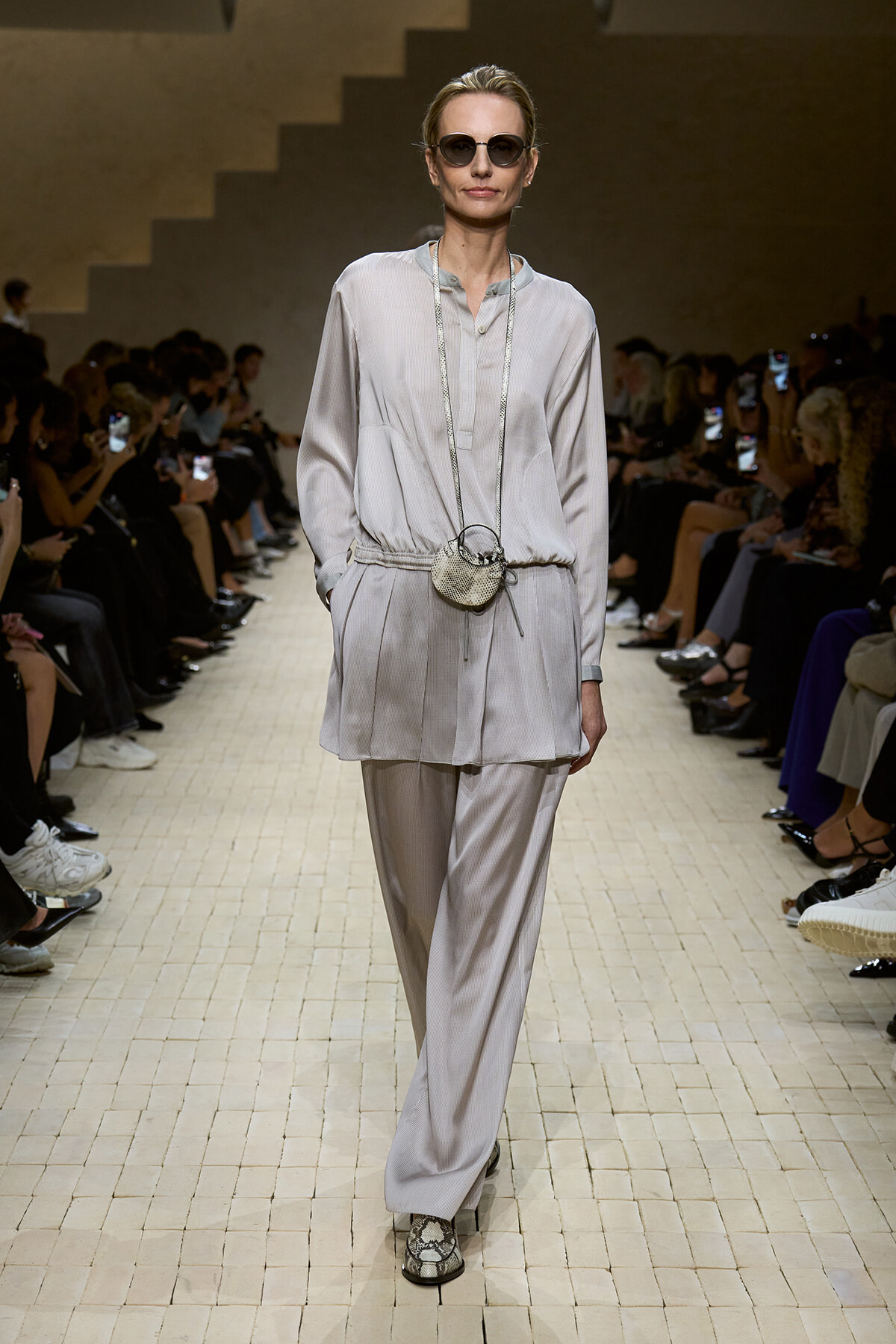 Model on a fashion runway wearing a pale gray tunic and wide-leg pants, round sunglasses, and a small silver circular crossbody bag.