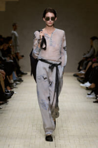 Model walks a fashion runway in a pale floral-pattern blouse and matching wide-leg pants, with a black fringe belt and sunglasses, carrying a black bag on her shoulder.